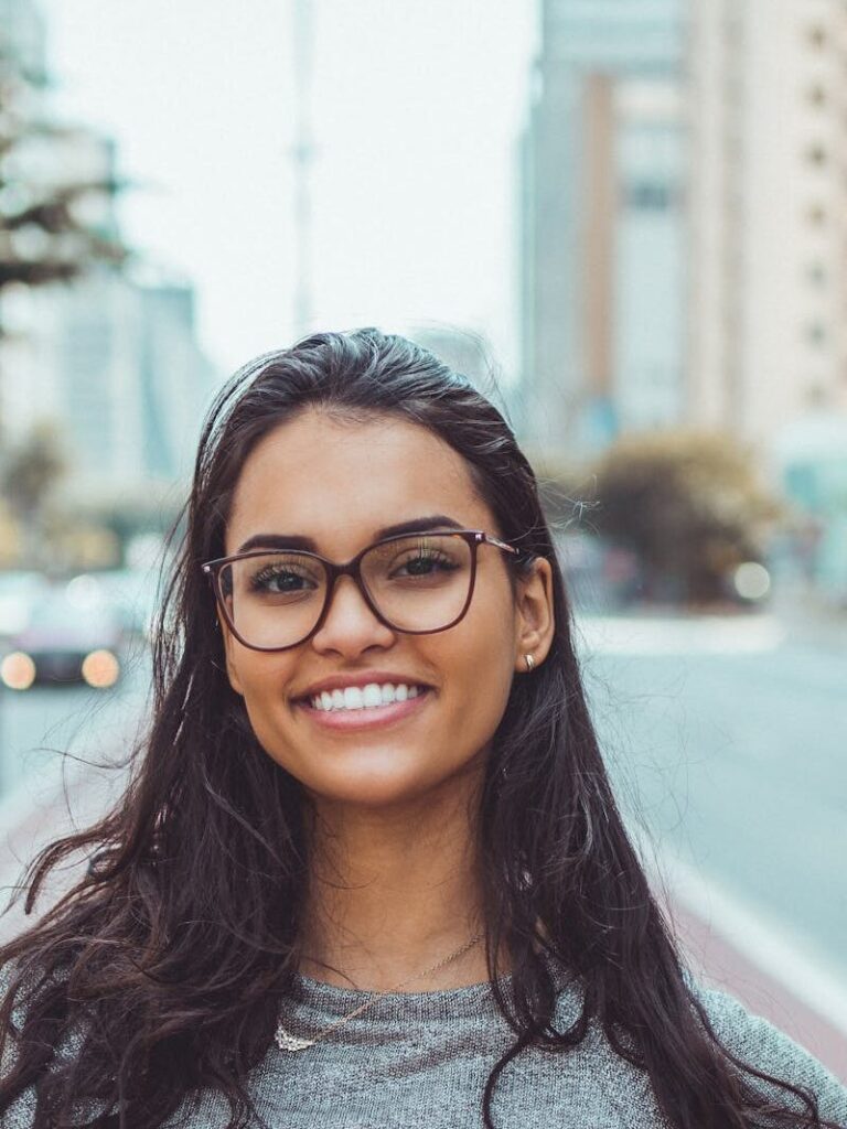 A young woman with glasses smiling on a city street, embracing urban lifestyle.